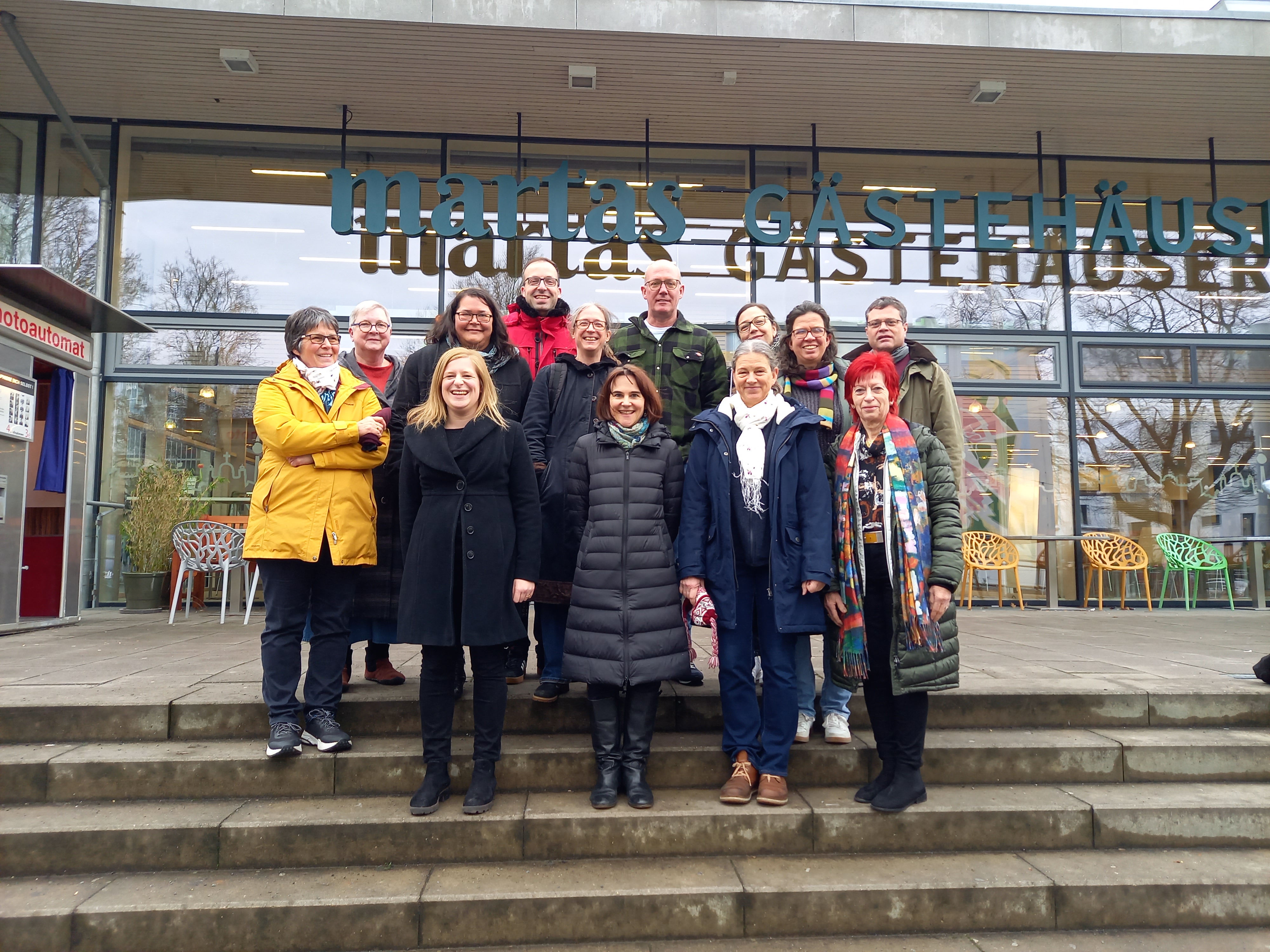Gruppenbild des BackStage Teams: 13 Personen stehen auf einer Treppe vor einem Gebäude.