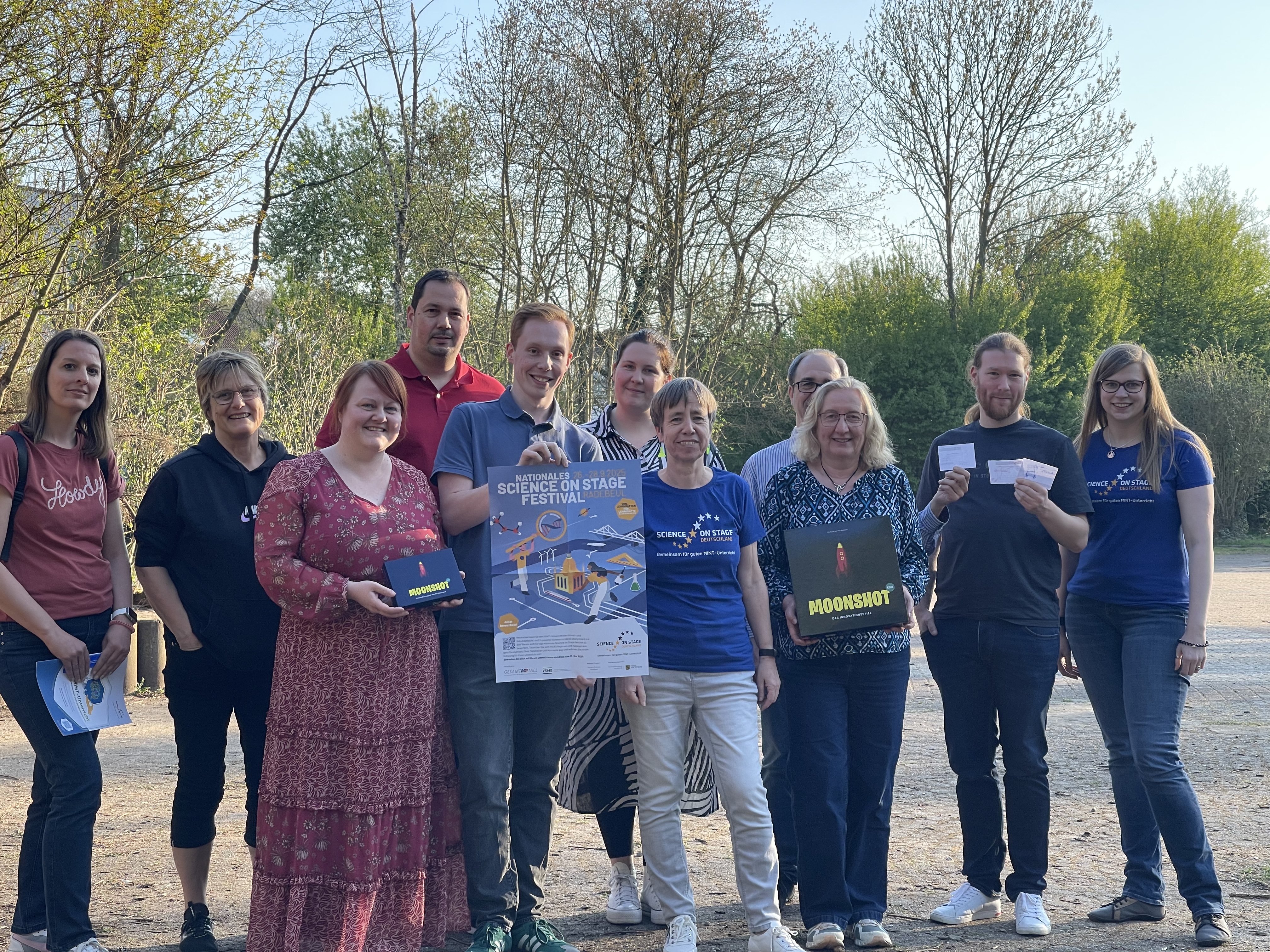 Elf Personen stehen draußen auf einer Freifläche vor Bäumen. Eine Person hält ein Poster vom Nationalen Science on Stage Festival. Weitere Personen halten Teile des Spiels Moonshot.