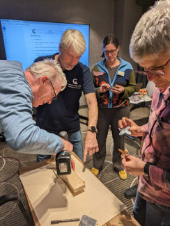vier Personen, ein Mann bohrt ein Loch in ein Stück Holz, das auf einem Tisch liegt