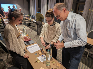 drei Personen basteln, auf einem Tisch liegen Stücke aus dünnem Blech und eine Zange