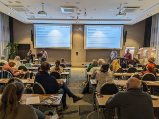 Gruppenfoto vom Plenarraum von hinten, vorne stehen vier Referenten