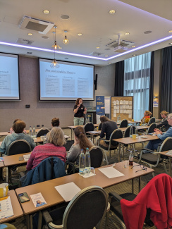 Gruppenfoto vom Plenarraum von hinten, vorne steht eine Referentin