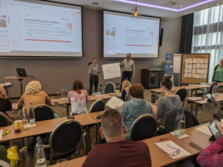 Gruppenfoto vom Plenarraum von hinten, vorne stehen zwei Personen mit einem Plakat